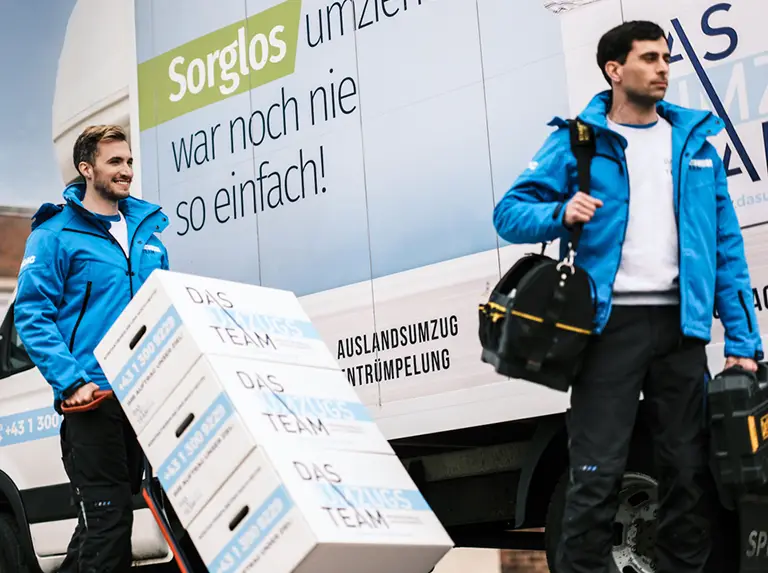 Zwei Umzugsmonteure von DasUmzugsteam gehen mit Umzugskartons und Handwerkskoffer bei dem Umzugs-LKW vorbei