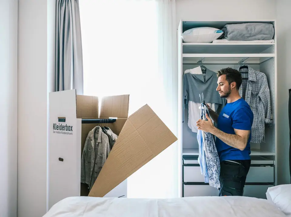 Umzugsmonteur packt Kleider aus Kleiderschrank sorgfältig in einen Umzugskarton.