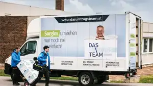 1 Möbelpacker mit einer Umzugsrodel und ein zweiter Möbelpacker stehen vor einem LKW