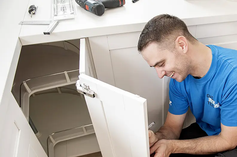 Hilfe beim Küchenaufbau: Ein junger Monteur in blauem T-Shirt kniet vor einem geöffneten Küchenschrank und hält einen Stift in der Hand.