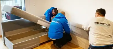 Wiener Möbelpacker: 3 Umzugshelfer knieen am Boden und demontieren ein graues Bett.